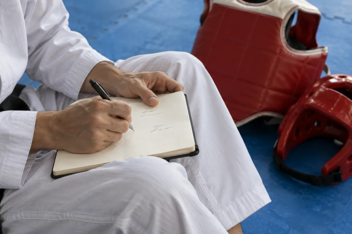 A taekwondo fighter in a white dobok writes in a notebook on their knee, with a red trunk protector and head guard on the mat beside them.